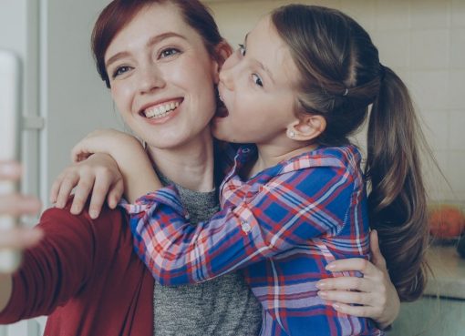 Smiling mother together with funny playful daughter making selfie photo with smartphone camera at home in kitchen. Family, cooking, and people concept
